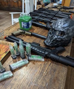 black airsoft guns lined up on a wooden counter with green pellets in a container and a black and grey protective mask