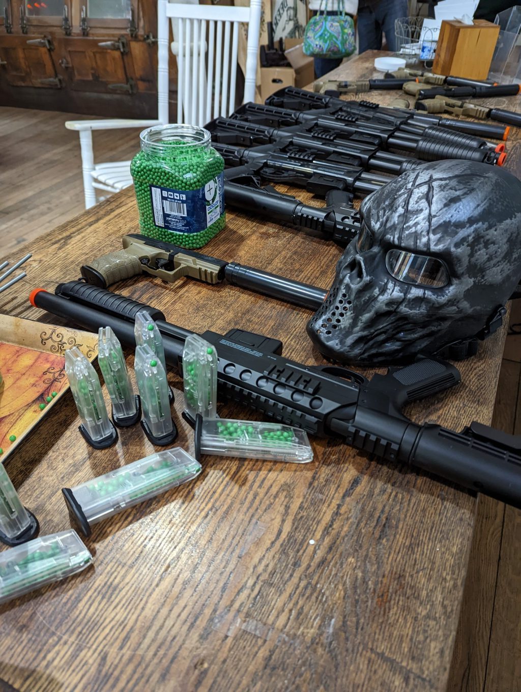 black airsoft guns lined up on a wooden counter with green pellets in a container and a black and grey protective mask