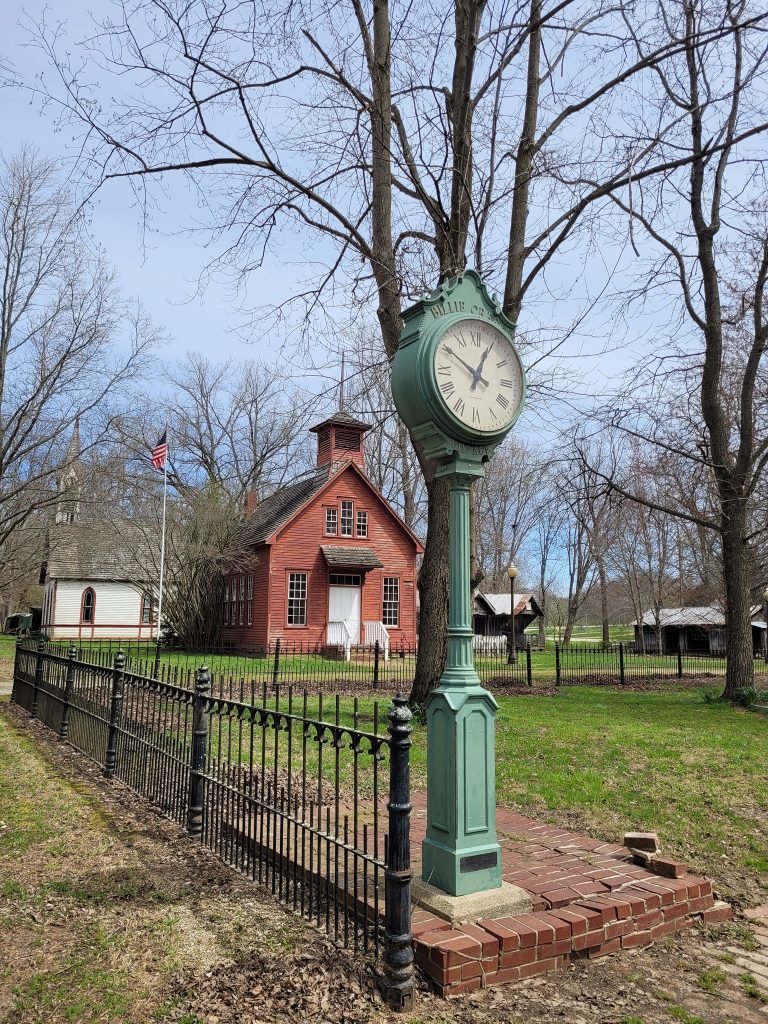 green clock with schoohouse in the background in a grassy area bare trees and black iron fence