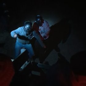 dark image with flashlight highlighting two men hunched over with airsoft masks on
